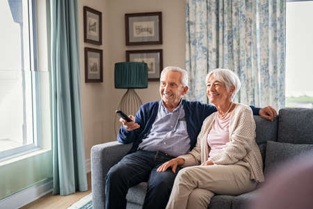 Happy senior couple in living room using remote control to change channel. Seniors watch TV for entertainment while man using remote control. Cheerful wife and old man sitting on couch in living room with television show.の写真素材