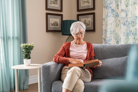 Old beautiful woman reading a book while sitting on couch at home. Elder lady relaxing at home while reading a book on sofa. Senior grandmother wearing eyeglasses for read a novel alone.の写真素材