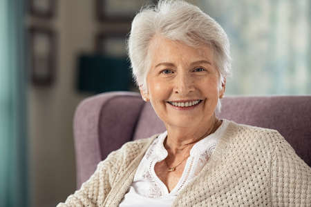 Portrait of joyful old woman sitting on armchair looking at camera. Retired elderly woman smiling while sitting alone in care centre. Portrait of an elegant senior grandmother relaxing at home with copy space.の写真素材