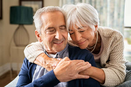 Romantic senior woman embracing from behind her husband sitting on sofa. Cute old couple in love hugging with closed eyes. Retired woman consoling her husband at home.の写真素材