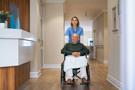 Happy nurse helping old man in wheelchair in nursing home. Young healthcare worker taking care of an elderly disabled patient at care facility centre. Friendly caregiver pushing old patient sitting on a wheelchair in care centre.の写真素材
