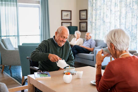 Smiling senior man pouring tea in cup from teapot in care facility with wife sitting at table in the common area. Elderly man in care centre sitting on wheelchair and serving tea. Serene disabled senior man enjoying tea time with friends during a visit at nursing home.の写真素材