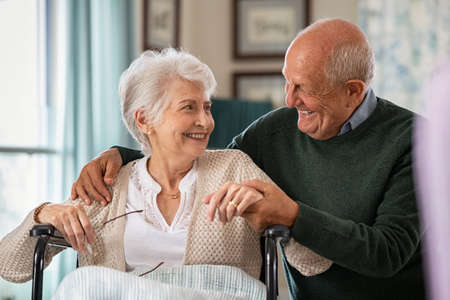 Romantic senior husband embracing disabled wife sitting on wheelchair at nursing home. Cheerful old couple embracing and looking at each other with love. Happy elderly man holding hands of handicapped woman sitting in wheelchair at care centre.の写真素材