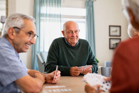Retired senior man in wheelchair playing cards with friends at nursing home. Smiling group of old people playing cards at care centre. Happy and content seniors playing at lunch table and havin fun together.の写真素材