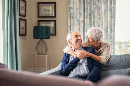 Happy senior woman embracing her husband at home while laughing together. Smiling wife hugging old man sitting on couch from behind. Joyful retired couple having fun at home while looking at each other with copy space. Love and unity concept.の写真素材
