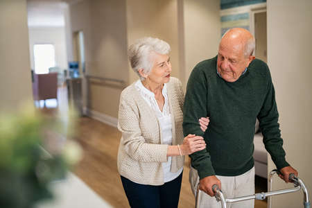 Elderly woman help her husband walking using walker in nursing home. Senior woman helping disabled man with walking frame. Lovely wife holding hands of old husband in care centre.の写真素材