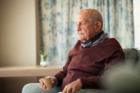 Depressed senior man sitting on armchair holding disposable cup of coffee and thinking. Frustrated retired man sitting on sofa and drinking a cup of hot tea. Sad mature man sitting alone at nursing home with sad expression.の写真素材