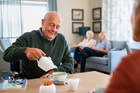 Smiling old man pouring hot tea in cup from kettle in nursing home. Elderly man in care centre sitting on wheelchair and drinking tea. Happy disabled elder in wheelchair enjoying tea time in the afternoon together with other patient of the care facility.の写真素材