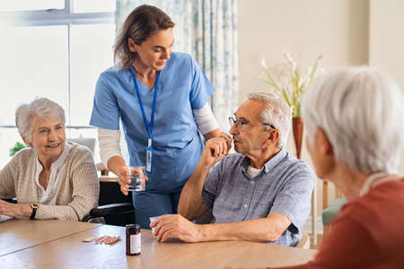 Young nurse in uniform giving medicine to group of seniors at modern nursing home. Caregiver giving medicine to old patient sitting at table at care facility centre. Elderly man taking pills from healthcare worker at retirement community together with other patients.の写真素材