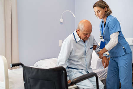 Lovely nurse assisting senior man to get up from bed at hospital. Caring nurse supporting elderly patient while getting up from bed and move towards wheelchair at nursing home. Disabled man standing up at care centre while caregiver helping him to get up in the morning.の写真素材