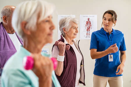 Group of old happy people using dumbbells for workout session with physiotherapist. Group of senior man and elderly woman exercising with dumbbells in clinic with trainer. Seniors doing workout weight lifting for wellness with young instructor.の写真素材