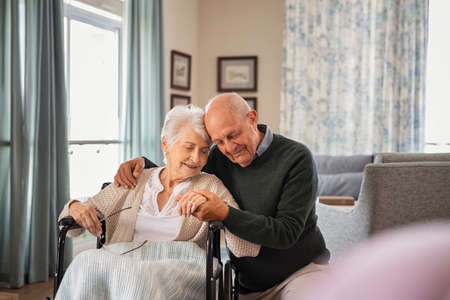 Romantic senior man embracing woman sitting on wheelchair at nursing home. Elderly couple in love embracing with eyes closed and deep respect for each other. Lovely old man holding hands of his disabled wife at care centre during a visit.の写真素材
