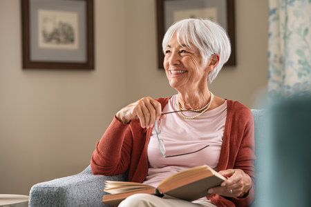 Cheerful senior woman holding book and eyeglasses thinking while relaxing at home. Happy elderly woman reading book at home sitting on couch. Beautiful old teacher takes a break from reading while looking through the window with a big grin.の写真素材
