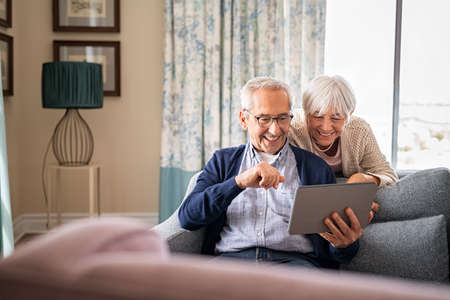Happy old couple sitting on sofa using digital tablet with copy space. Cheerful senior man and beautiful elderly woman relaxing at home while watching funny video on digital tablet. Smiling grandparents making a video call with their nephews during lockdown.の写真素材