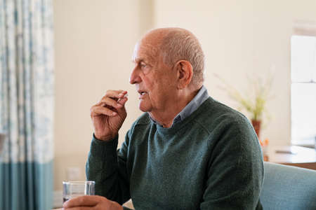 Ill senior man taking medicine at care centre. Elderly man taking pills for depression sitting on couch. Old upset patient swallowing pill with glass of water.の写真素材