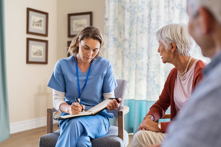 Nurse talking to old patients while being in a nursing home. Young healthcare worker visiting senior couple at care centre and writing information. Nurse wearing uniform noting symptoms of elderly woman.の写真素材