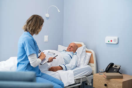 Nurse writing recovery information on clipboard with old patient lying in hospital bed. Young female doctor writing medical notes on clipboard with senior patient in care centre. Hospitalized man lying on bed while nurse checking his status.の写真素材
