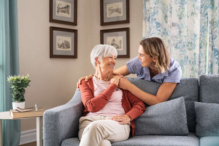 Smiling adult daughter embracing from behind old mother sitting on sofa and laughing together. Excited aged woman having fun with mature daughter during home visit. Old mom feeling happy with adult daughter while looking at each other.の写真素材