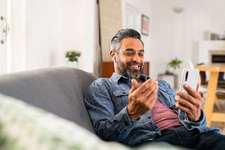 Happy smiling indian man using smartphone to do a video call sitting on sofa. Mid adult indian man on couch talking to his colleague or friend in video conference while using wireless earbuds. Mature mixed race man relaxing at home using cellphone during videocall, copy space.の写真素材
