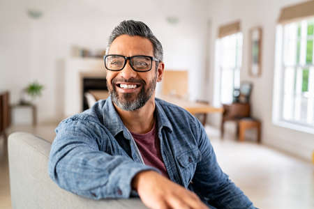 Happy mature middle eastern man wearing eyeglasses with beard sitting on couch at home. Portrait of indian man relaxing at home and looking away with big smile. Handsome mid adult guy with specs thinking about his future.の写真素材