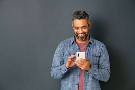 Mid adult multiethnic man texting phone message on smart phone isolated on grey background. Smiling middle eastern man using smartphone leaning on gray wall. Happy mixed race guy using new app on mobile phone with copy space.の写真素材