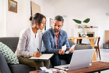 Mature couple calculating bills at home using laptop and calculator. Multiethnic couple working on computer while calculating finances sitting on couch. Mature indian man with african american woman at home analyzing their finance with documents.の写真素材