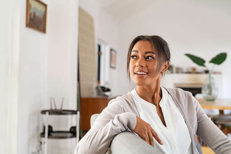 Thoughtful mature african woman sitting on couch at home and looking outside the window. Contemplative beautiful woman relaxing on sofa in the living room. Smiling mid adult woman with copy space contemplating.の写真素材