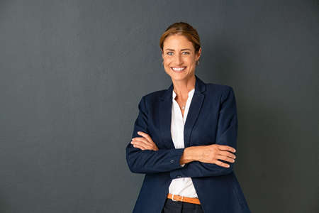 Confident mature woman with crossed arms in formal clothing with copy space looking at camera. Happy smiling mid adult woman standing against grey background. Portrait of beautiful positive businesswoman.の写真素材