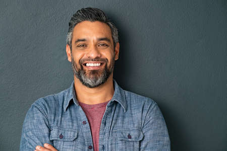 Close up face of handsome mature man with beard looking at camera. Happy mid adult indian man isolated against grey wall. Portrait of smiling mixed race guy on gray background with copy space.の写真素材