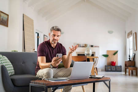 Mature businessman on video call with wireless earphone. Mid adult man working from home during online meeting with smartphone. Entrepreneur sitting on couch doing conference over smart phone during lockdown.の写真素材