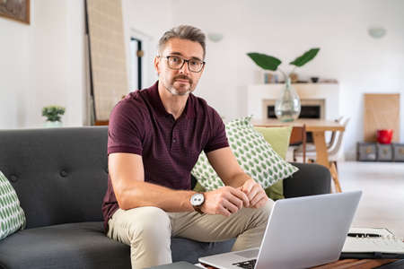 Portrait of mature man wearing spectacles and working from home. Handsome mid adult man sitting on sofa while working on laptop and looking at camera. Businessman using laptop in telecommuting from home: telecommuting and smart work concept.の写真素材