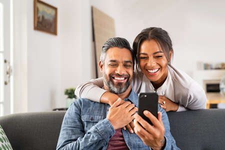Mature multiethnic couple at home using smart phone together and smiling. Mid adult man relaxing on sofa and showing new app to african american wife on cellphone. Middle eastern man and woman sitting on couch at home and using mobile phone to do a video call with family or friends.の写真素材