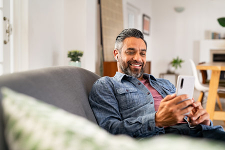 Happy mid adult man using smartphone device while sitting on sofa at home. Smiling indian man lying on couch reading messages on mobile phone while listening to music with wireless earbuds. Mature guy relaxing at home.の写真素材
