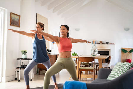 Two mature women doing warrior pose at home with stretched arms. Healthy mature woman doing yoga exercise with mixed race friend in living room. Middle aged lady in sportswear exercising together at home.の写真素材