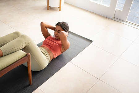 Top view of mid adult fit woman doing press on a mat while keeping legs on chair at home. African american woman practicing crunches at home during morning routine. High angle view of healthy mixed race girl doing abs exercise and strengthen her muscles.の写真素材