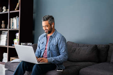 Successful mature indian businessman sitting on couch typing on laptop with wireless earphones. Mixed race businessman sitting on couch while working from home during video call. Happy multiethnic business man in casual clothing working on computer.の写真素材