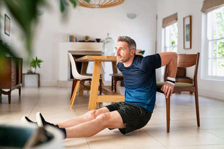 Mature man doing fitness exercise while doing pushups using chair in living room. Middle aged man training at home wearing sportswear during fitness exercises for triceps. Mid adult fit guy exercising for wellness in his living room.の写真素材