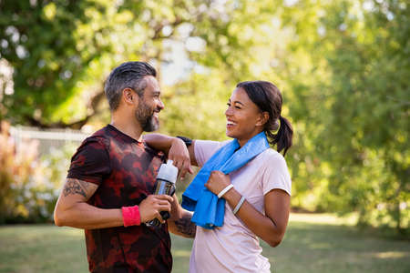 Cheerful multiethnic couple resting together after jogging in the park. Happy mature man and beautiful woman laughing while resting after running during summer afternoon. Smiling woman and carefree indian man taking a break after fitness training workout while looking at each other.の写真素材