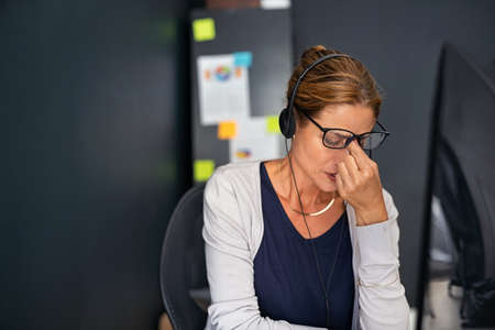 Upset businesswoman wearing eyeglasses with headset suffering from headache while working from home. Tired mature woman wearing headphone suffering from migraine after conversation with a client. Middle aged business woman feeling unhappy working from home: overworked and burnout concept.の写真素材