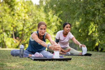 Two mature woman stretching their legs after exercise in park. Beautiful athletic woman doing stretches in the park with her mixed race friend. Sporty mid adult lady stretching after a good workout session outdoor.の写真素材