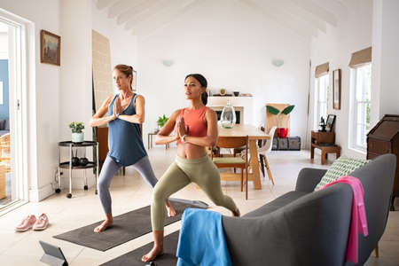 Beautiful mature woman friends practicing yoga at home. Sporty fit african american woman practices yoga asana warrior pose with folded hands at home with her friend. Two mid adult lady practicing watching online sport lesson on digital tablet.の写真素材