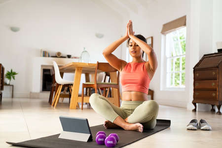 Mature woman in sportswear sitting on yoga mat and looking at digital tablet for online lessons of hata yoga. Healthy mid adult black woman practicing yoga with online trainer at home. African american healthy lady practicing yoga while watching video tutorial.の写真素材