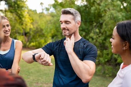 Mature tired man checking pulse after workout. Handsome middle aged man standing with group of friends measuring heart rate pulse on his neck and looking sport watch. Guy times the pulsations at park.の写真素材
