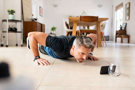 Mature man practicing pushups exercise while watching video on smartphone at home. Middle aged man doing fitness pushup workout at home while watching tutorial on mobile phone. Mid adult muscular guy working out doing plank.の写真素材