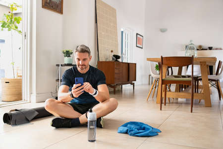 Mature handsome man using smart phone while exercising at home. Mid adult fitness man check his heart rate after daily workout on smartphone at home. Fit middle aged guy resting after strenght workout while using mobile phone.の写真素材