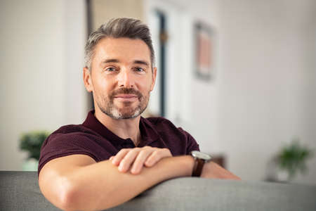 Portrait of handsome mature man relaxing on couch at home while looking at camera. Confident mid adult man resting while sitting on sofa in living room with copy space. Reliable businessman looking at camera with satisfaction.の写真素材