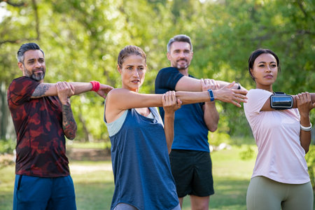 Mature woman and middle aged man stretching at park. Mid adult multiethnic group working out in park doing stretching exercise. Healthy group of mixed race friends doing stretching exercises together.の写真素材