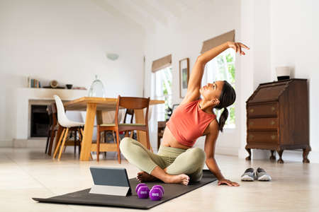 Mature mixed race woman in sportswear sitting on yoga mat doing stretching exercise while following online gymnastics class on digital tablet. Healthy african woman practicing yoga with online trainer on laptop at home with dumbbells. Fitness lady doing yoga exercise at home for healthy daily lifestyle.の写真素材