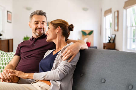 Mid adult couple embracing and relaxing on couch at home while holding hands. Romantic husband and beautiful wife hugging and looking at each other on sofa. Middle aged man relaxing at home with smiling woman.の写真素材