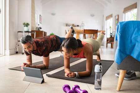Multiethnic mature couple exercising at home and watching training videos on digital tablet. Middle aged woman and indian man doing planks with a leg outstretched while watching fitness lessons online on digital tablet. Fit mid adult couple doing strenght plank following online tutorials.の写真素材